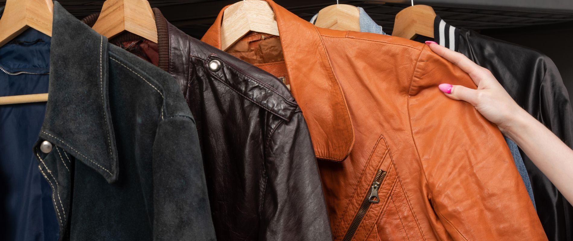 Assorted leather jackets on wooden hangers, including brown, black, and orange, being browsed by a person with pink nail polish.