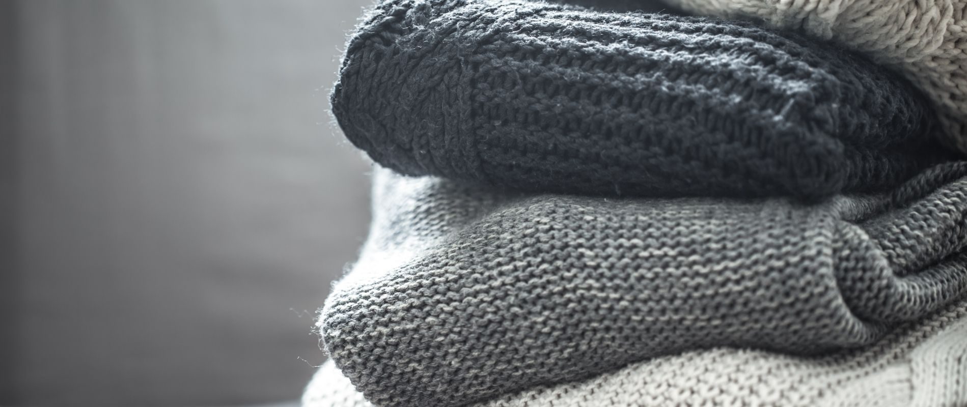 Stack of folded knitted sweaters in black, grey, and beige shades, showcasing cozy winter wear and texture detail.