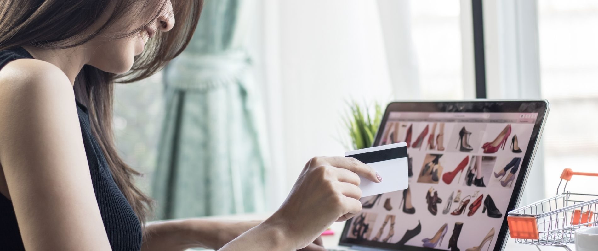 Woman shopping for shoes online with a credit card, laptop screen displaying various high heels, mini shopping cart nearby.