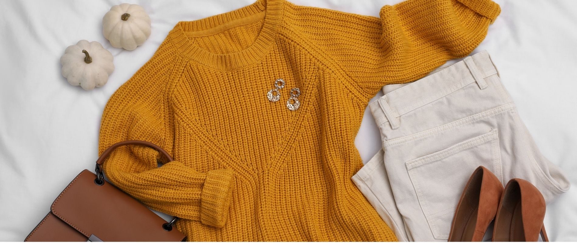 Flat lay of a fall outfit: mustard sweater, beige pants, brown high heels, brown handbag, earrings, and white mini pumpkins.