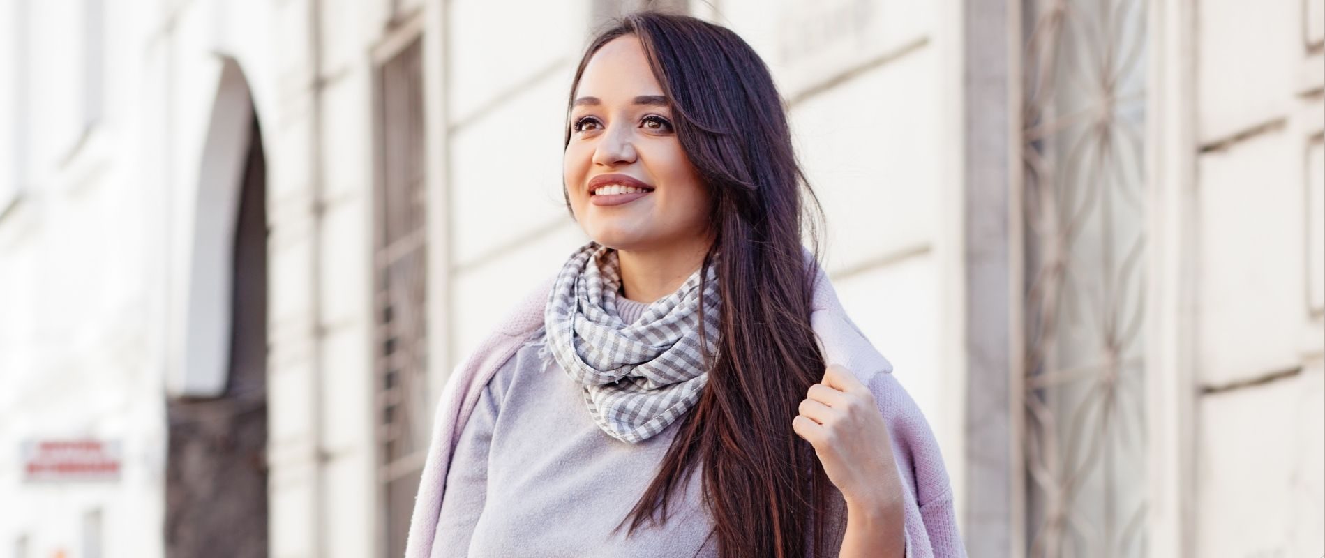 Smiling woman with long hair wearing a scarf and jacket, standing outdoors near a building on a sunny day.