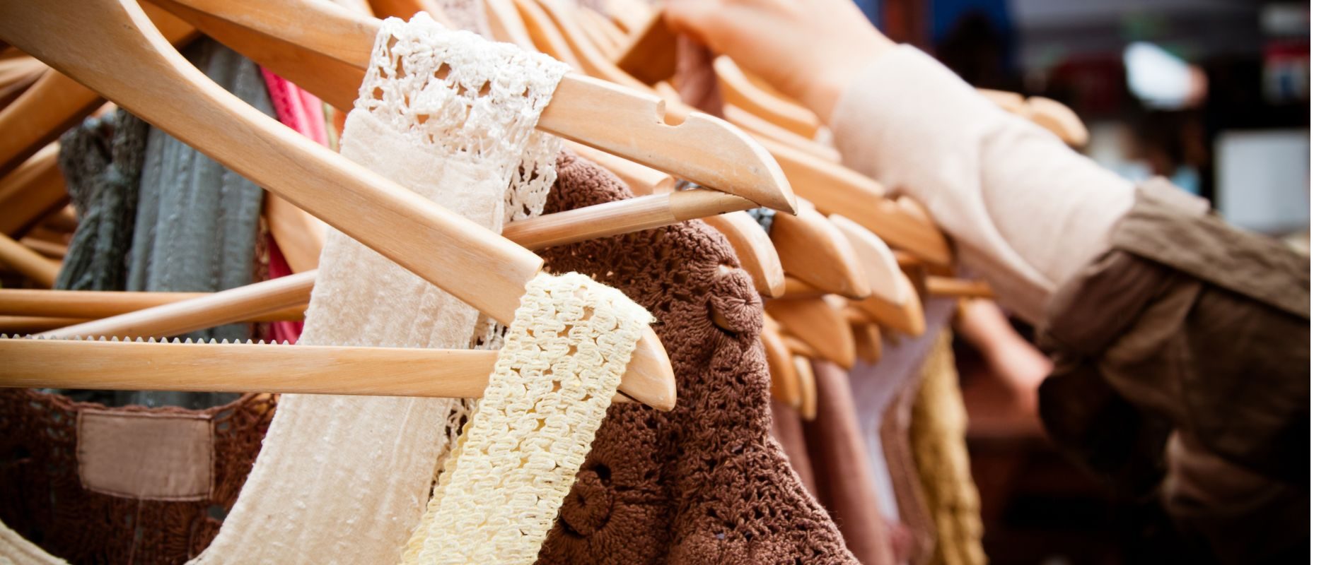 A person browsing hanging clothes in a store, featuring crocheted and knitted fabrics in various colors on wooden hangers.