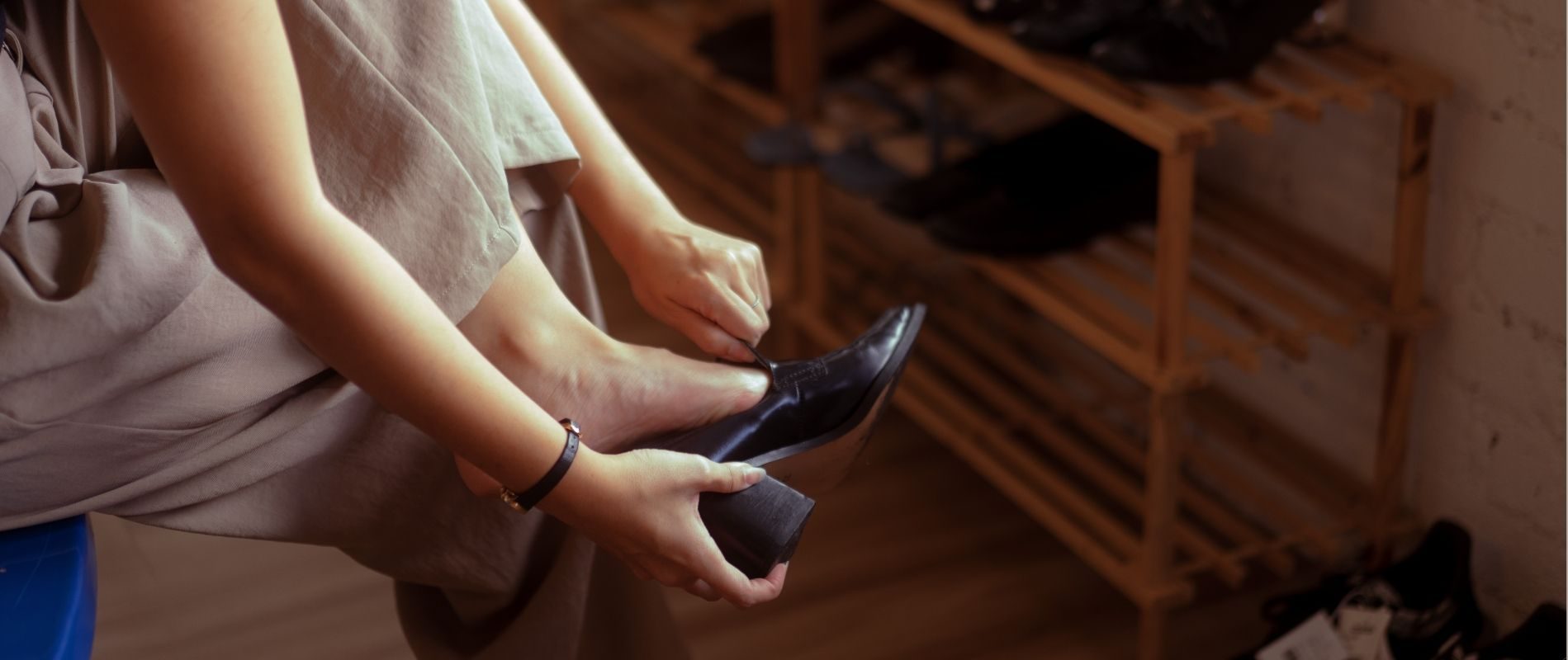 Person adjusting black high-heel shoe in a cozy setting with wooden shoe rack in the background.