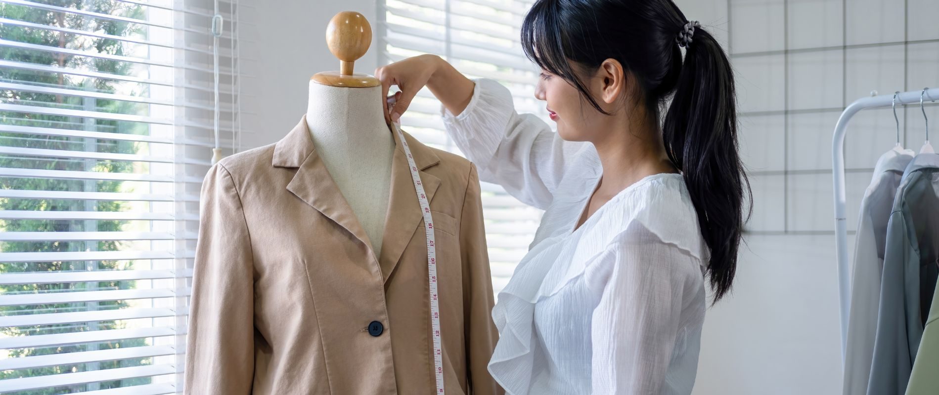 Seamstress measuring a beige blazer on a mannequin with a tape measure in a well-lit tailoring studio.