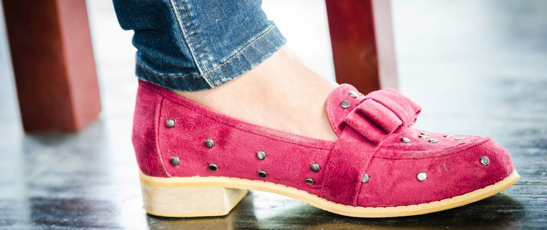 Close-up of a red suede flat shoe with metallic studs worn with blue jeans on a polished floor.