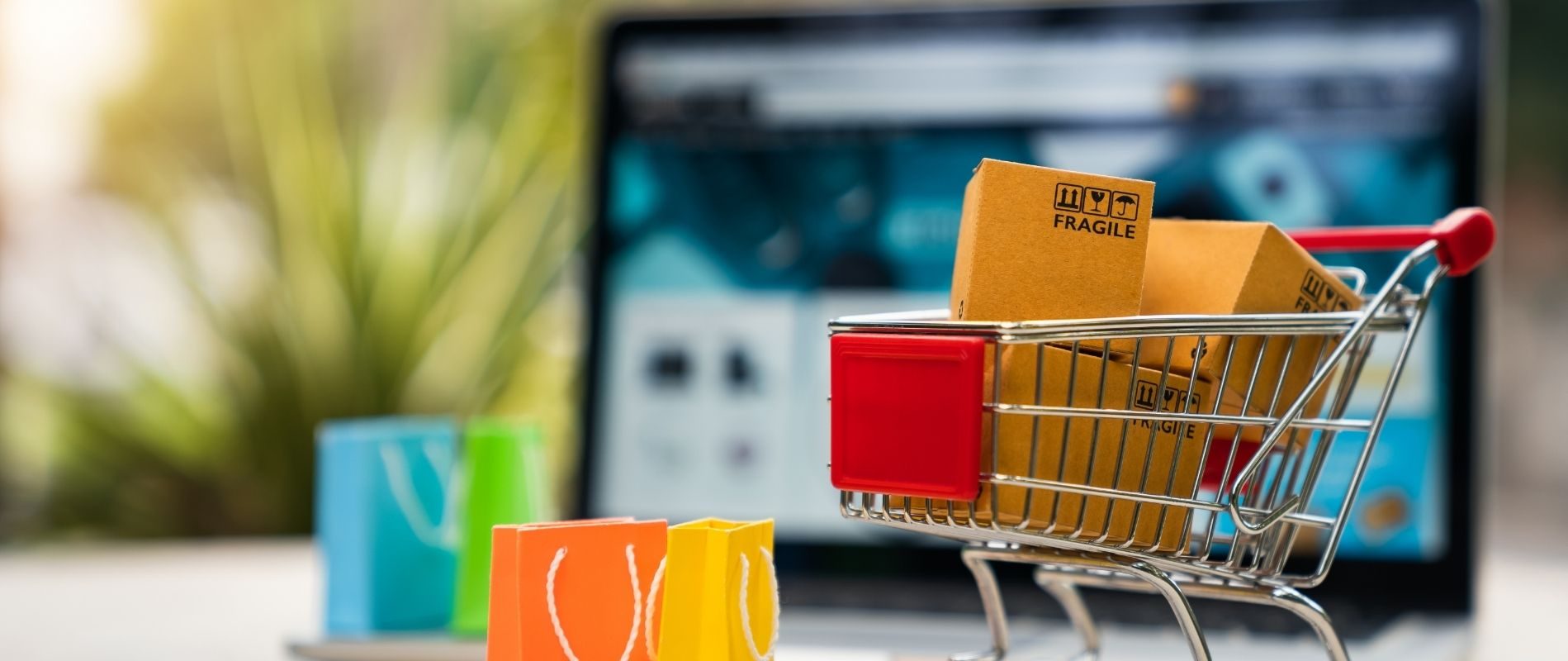 Shopping cart with boxes and colorful bags, suggesting online shopping in front of a laptop. E-commerce and online retail concept.