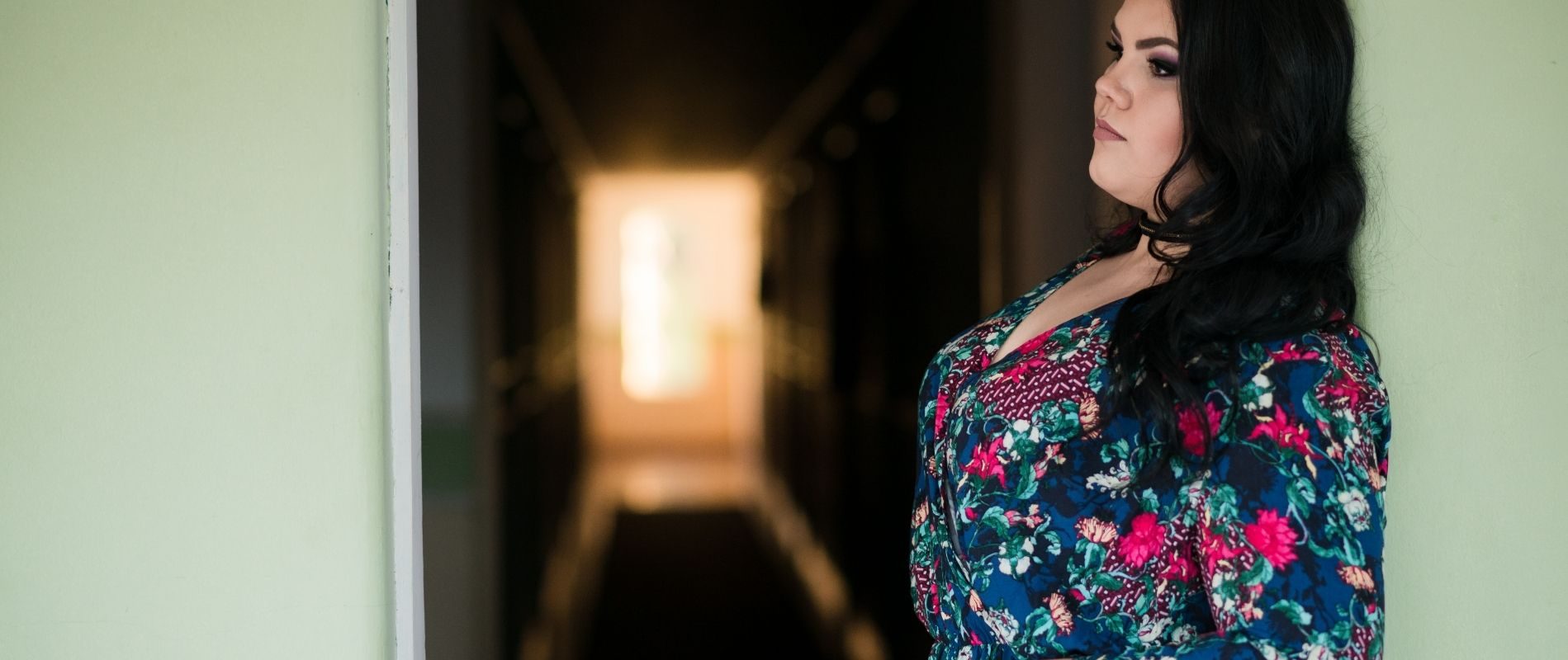 A person in a floral dress leans against a wall in a dimly lit corridor, illuminated by light from the far end.