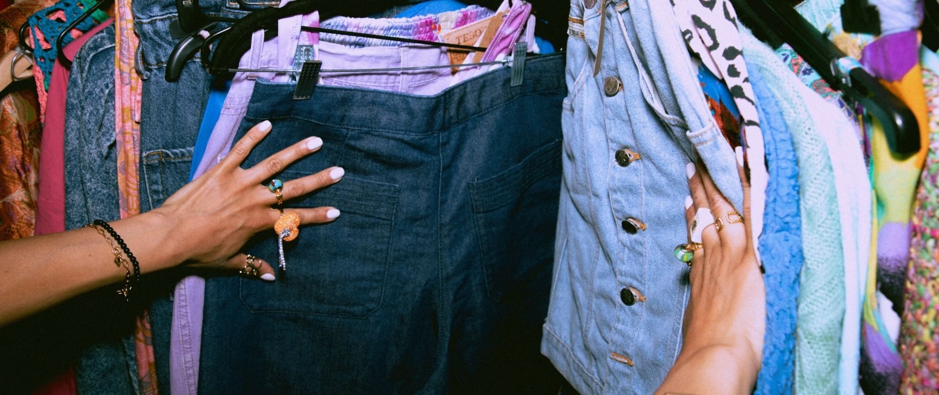 A person browses through various colorful clothes on a rack, showcasing denim shorts, patterned fabrics, and unique rings on their hands.