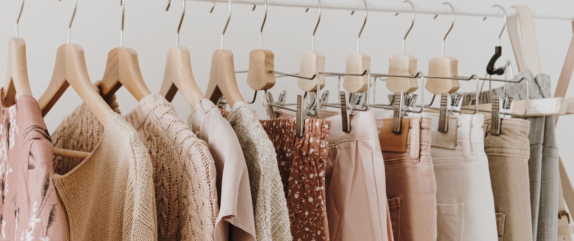 Assorted pastel-colored clothes on wooden hangers, including sweaters, pants, and shirts, neatly lined up in a wardrobe.