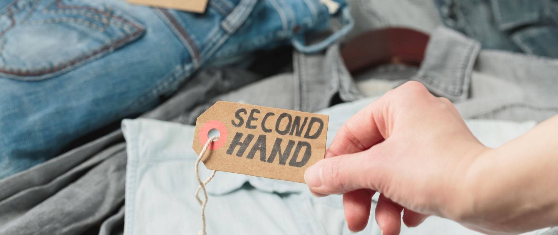 A person holds a "Second Hand" tag over a collection of folded clothing, including jeans and shirts, suggesting thrift shopping or resale.