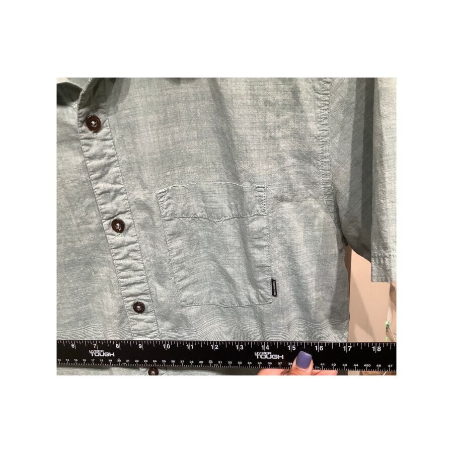 A person holds a ruler next to a green button-up shirt with a front pocket, likely measuring its dimensions. The ruler reads "TOUGH."