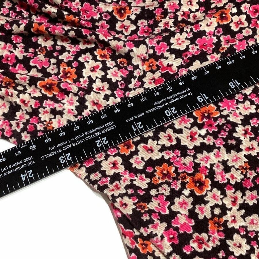 A black ruler measures floral-patterned fabric featuring pink, orange, and white flowers against a dark background. The fabric extends slightly wrinkled beneath the ruler.