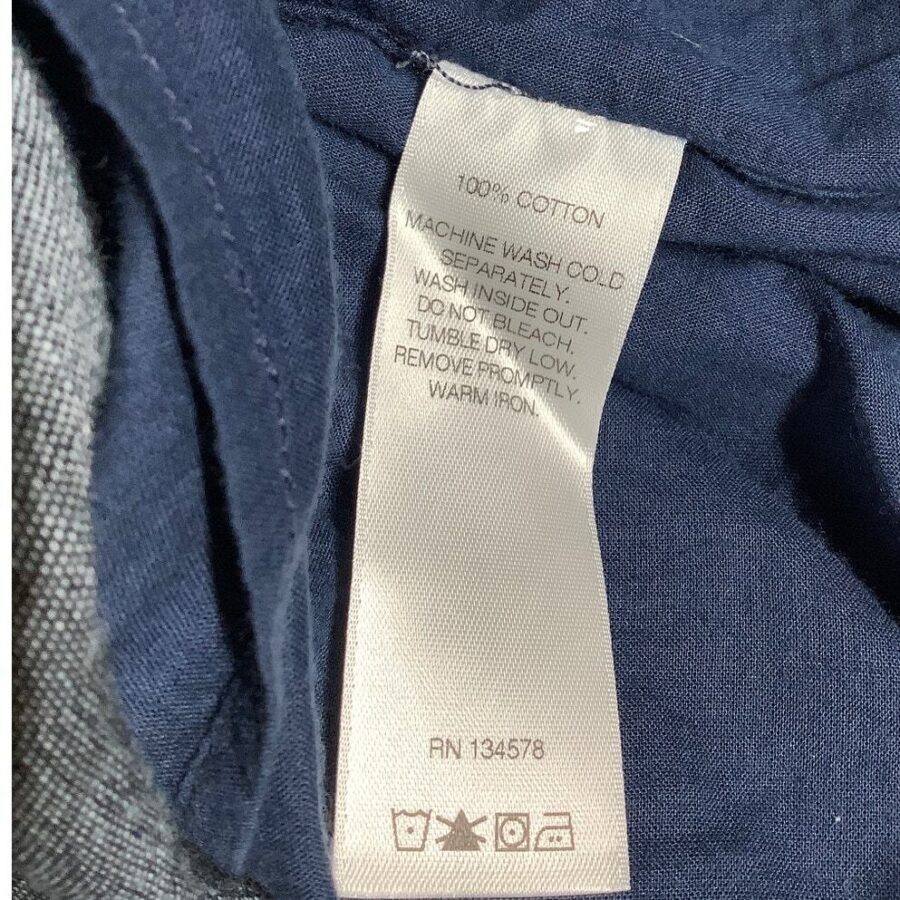 A clothing tag sewn into a dark fabric provides care instructions, specifying 100% cotton material. It advises cold washing, no bleach, and low tumble drying.