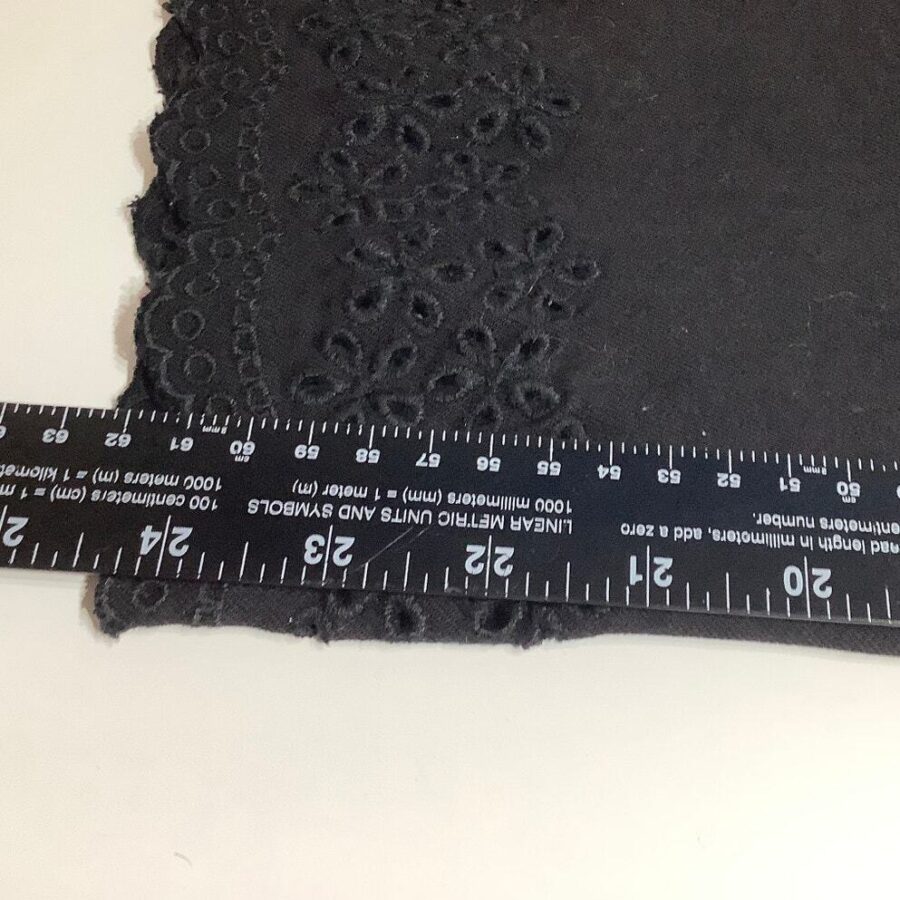 A black lace fabric with embroidered patterns is laid on a surface, measured with a ruler displaying both centimeters and inches.