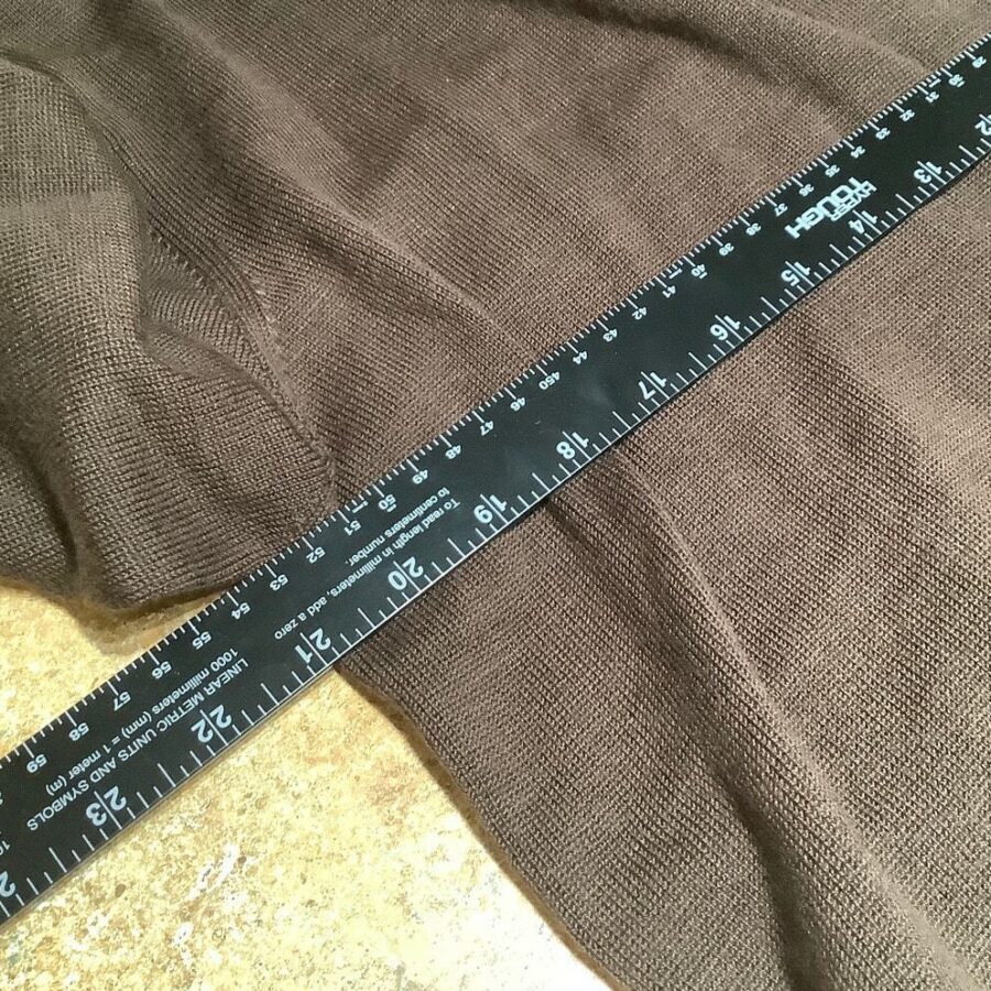 A black ruler with white markings is placed on a brown knit fabric laid on a textured surface. The fabric appears soft and cozy.