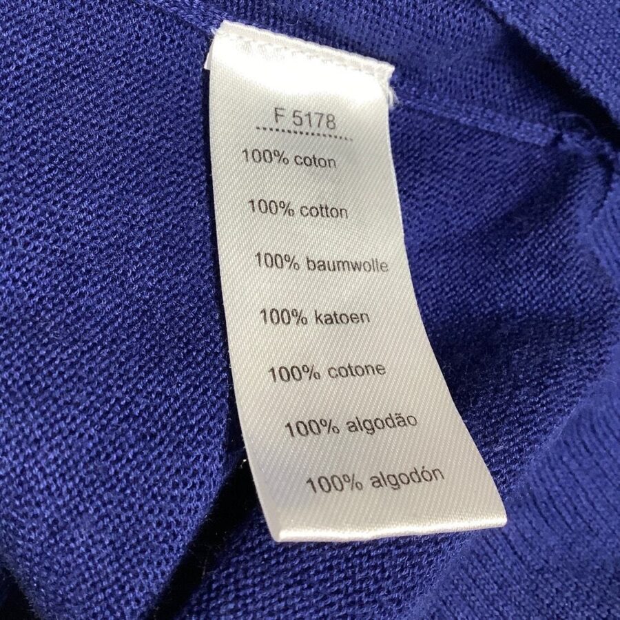 A white clothing label attached to a blue fabric, listing the material as 100% cotton in multiple languages. No landmarks or historical buildings present.