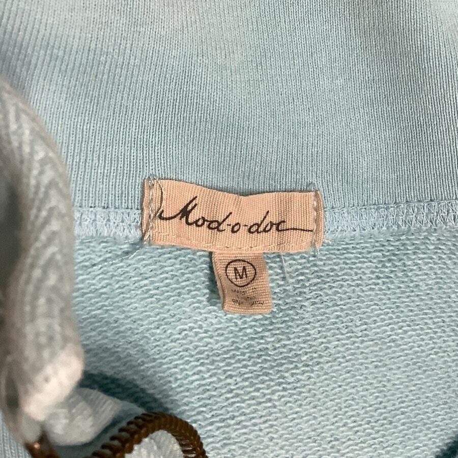 A close-up image of a light blue garment shows a fabric label with the text "Moda de" and a medium size indicator "M".