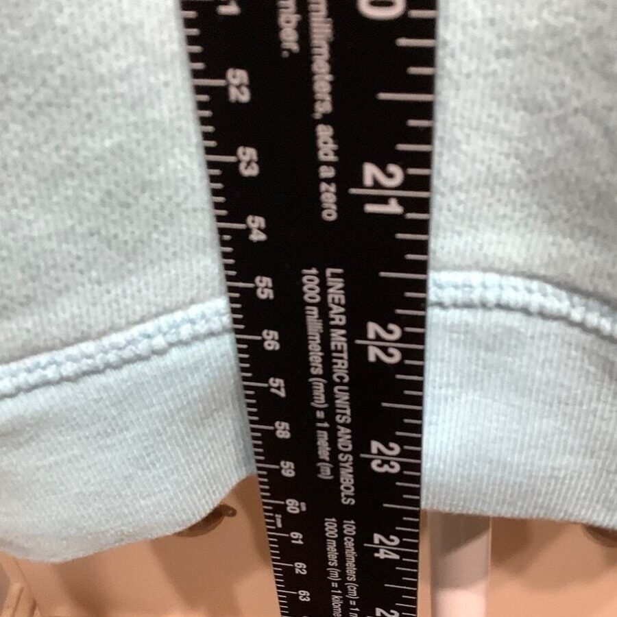 A black ruler with white markings is measuring the sleeve of a light blue garment. The ruler features both linear metric units and symbols.