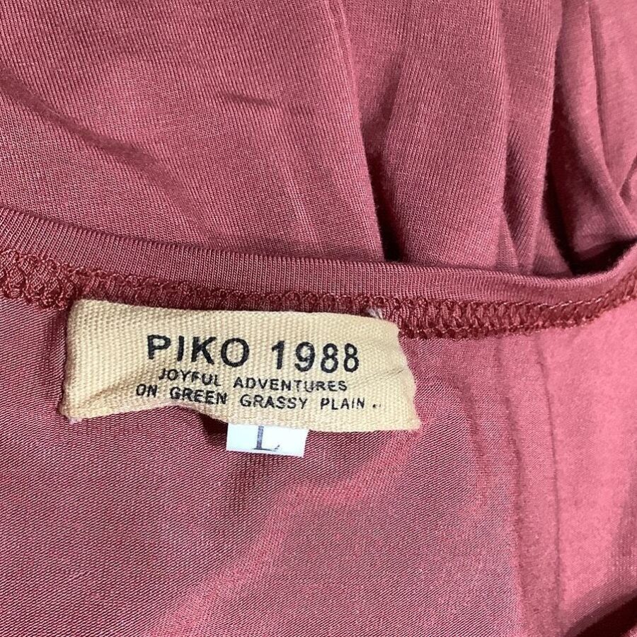 The image shows a close-up of a maroon garment with a label "PIKO 1988: Joyful Adventures on Green Grassy Plain" and an "L" size tag.