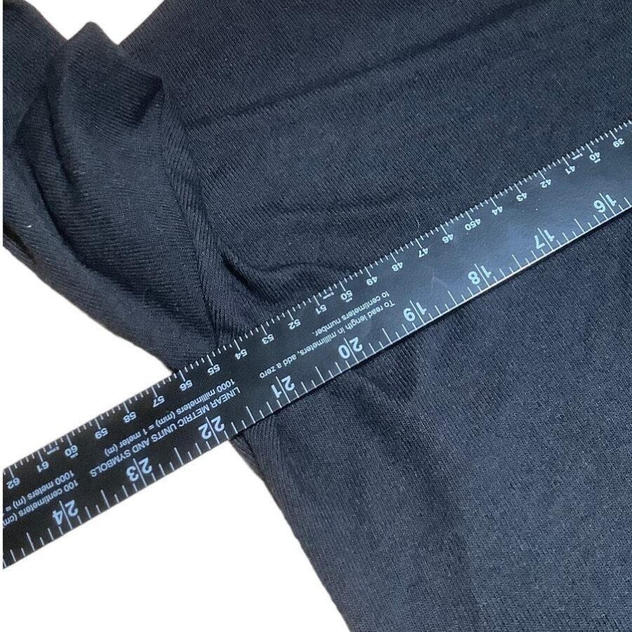 A dark fabric is measured with a black ruler, showing inch and centimeter markings, against a plain background. No landmarks or historical buildings are present.