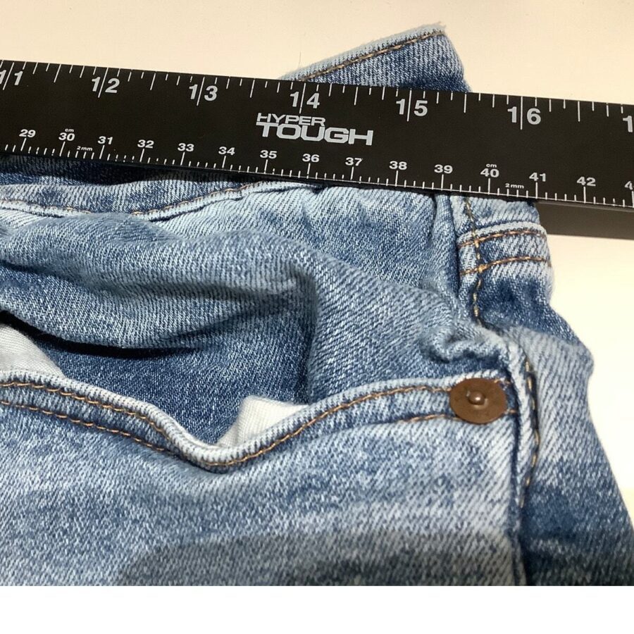 A pair of blue jeans measured with a black 12-inch ruler on a white surface, showing detailed stitching and a button.