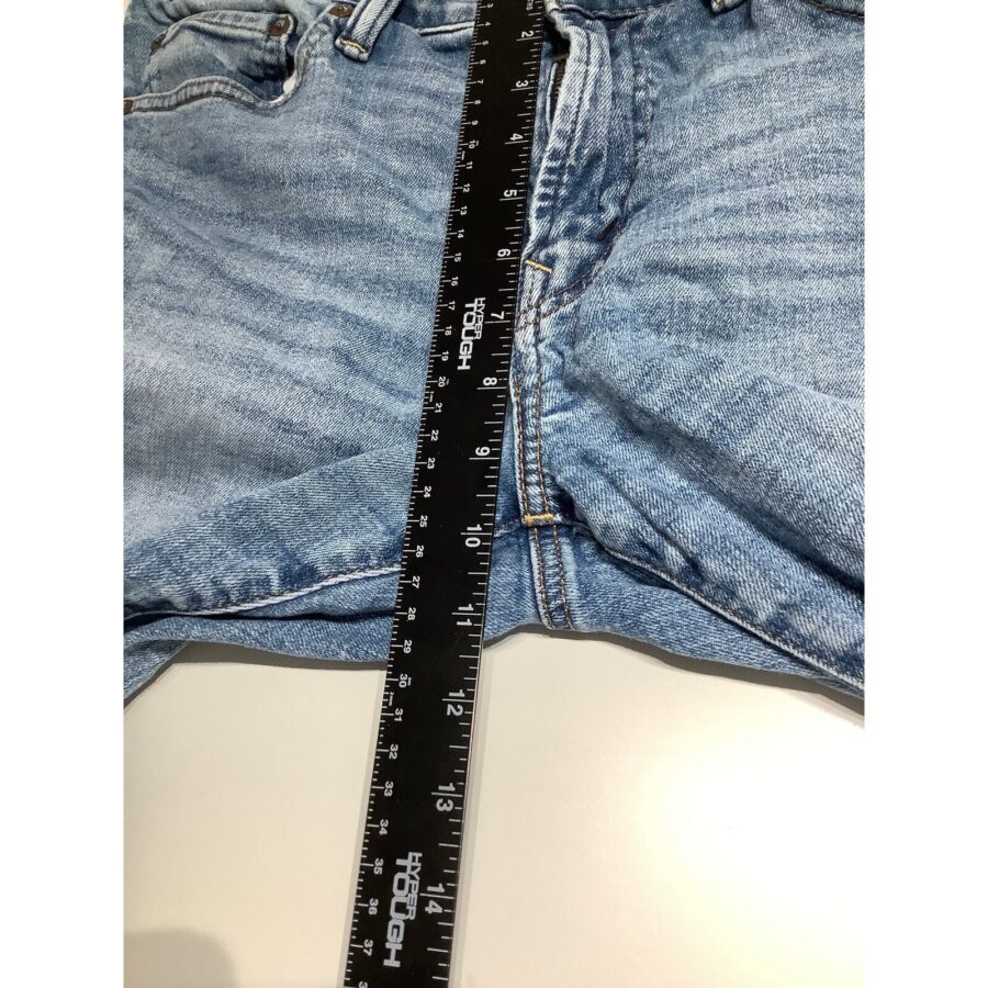 The image shows blue jeans laid flat on a surface with a black ruler measuring the inseam. No landmarks or historical buildings are visible.