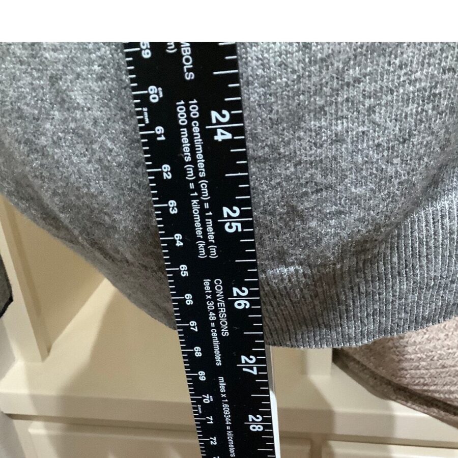 A black measuring tape with length conversions is held against a gray knitted garment. The tape shows measurements from 24 to 27 inches.