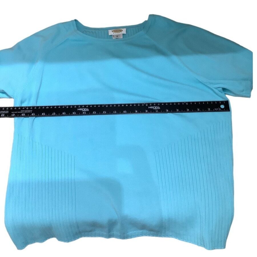 A blue short-sleeve sweater is laid flat with a measuring tape across it, showing the dimension of its width. No recognizable landmarks or buildings.