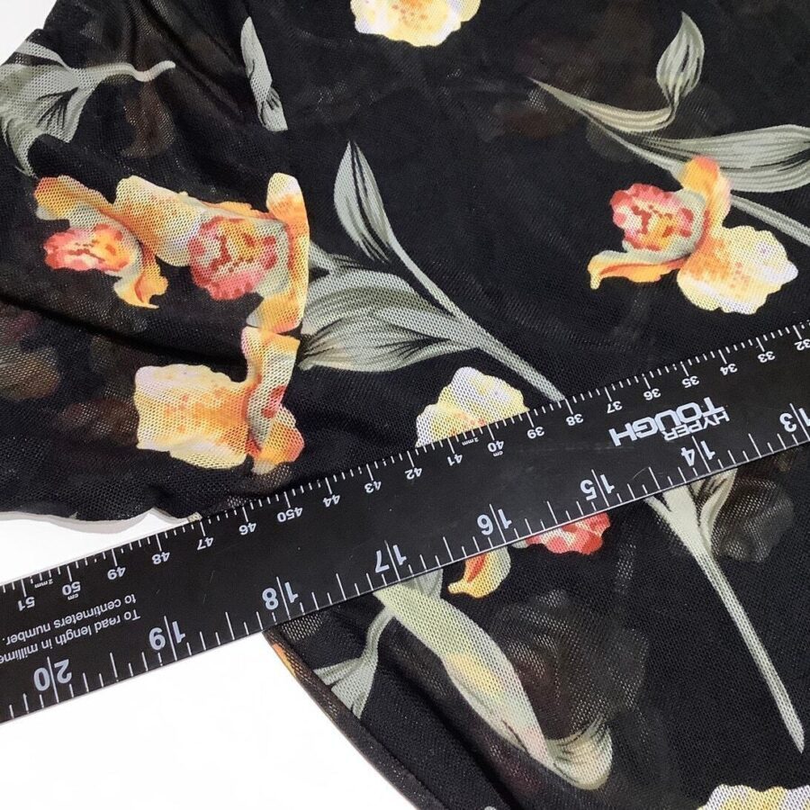 A black fabric with a floral pattern in orange, yellow, and green is measured with a black ruler displaying both inches and centimeters.