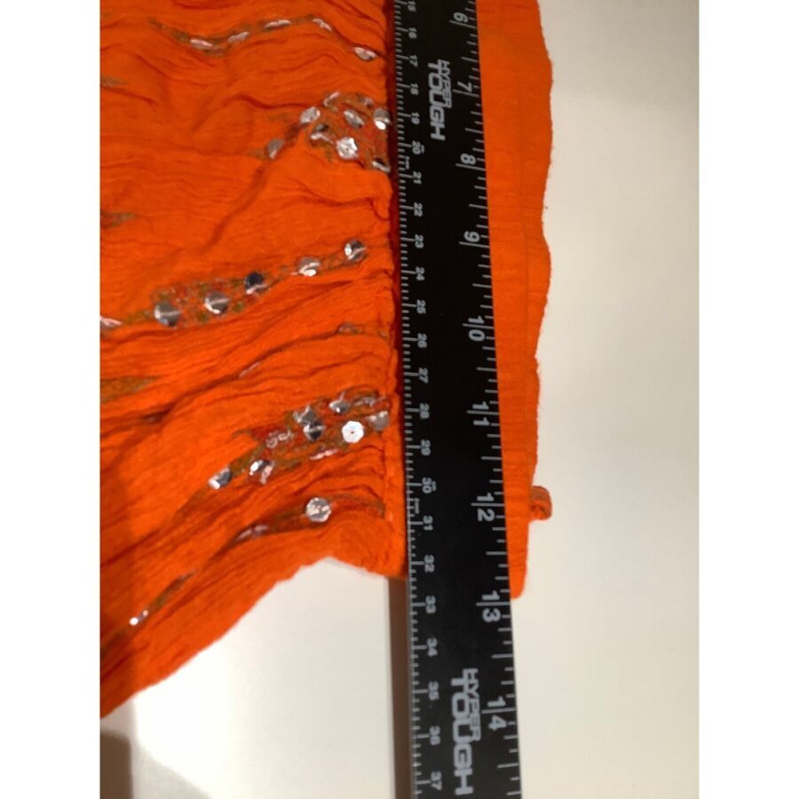 An orange fabric with sequins is being measured using a black and white ruler marked with inches and centimeters on a flat white surface.