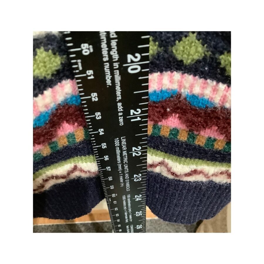 A close-up of a person holding a measuring tape against a colorful, patterned knitted fabric, showing a length measurement in inches and centimeters.