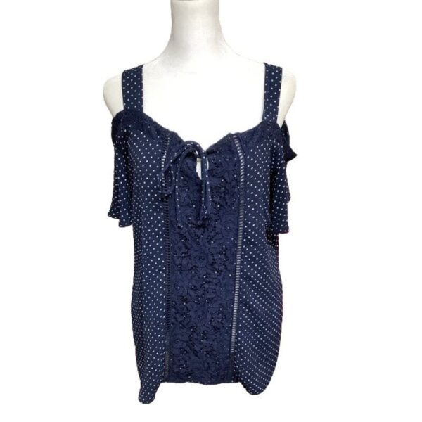 A dark blue, polka-dotted, sleeveless top with ruffle details and tie front, displayed on a white mannequin torso. No recognizable landmarks or historical buildings.