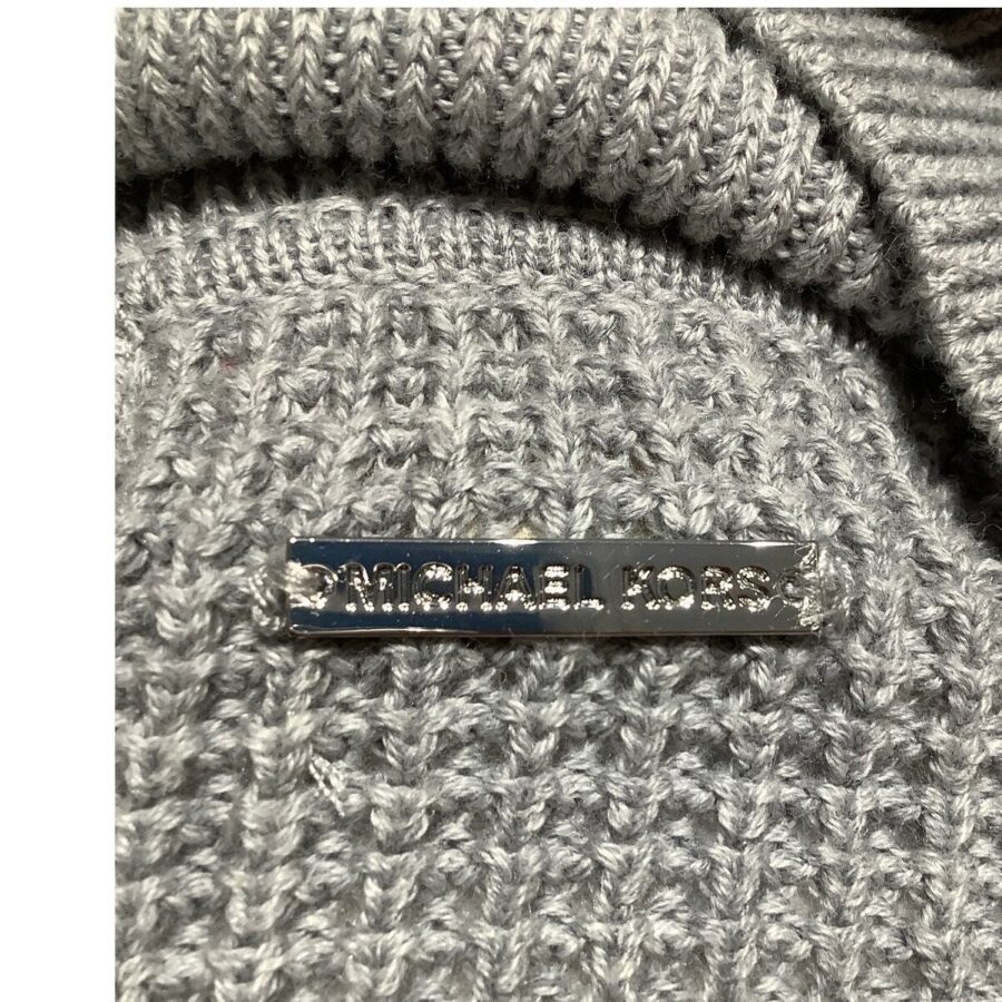 Close-up of a gray knitted fabric featuring a metallic label with "Michael Kors" engraved on it, positioned in the bottom left corner.