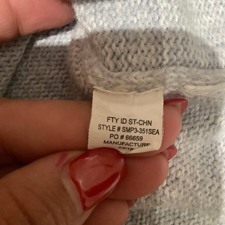 A person with red-painted nails holds a clothing tag displaying manufacturing and style information against a light blue knitted fabric.