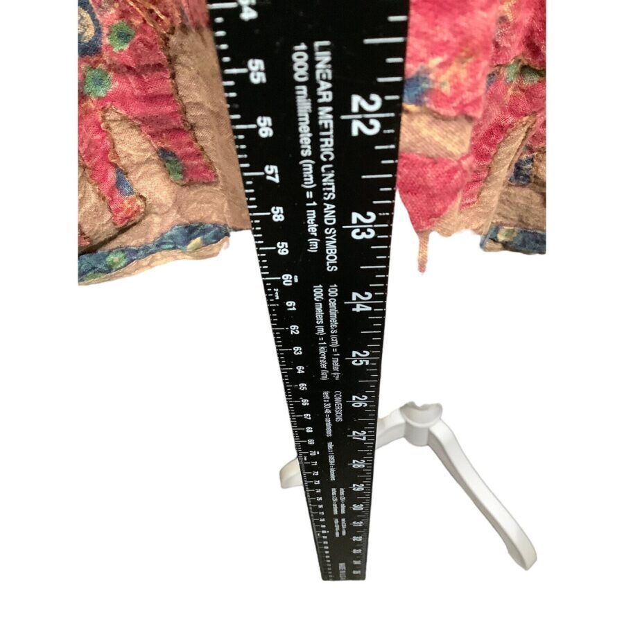 A metal measuring ruler leans against an ornate textile. The ruler reaches a white tripod stand, highlighting measurement details in millimeters and inches.