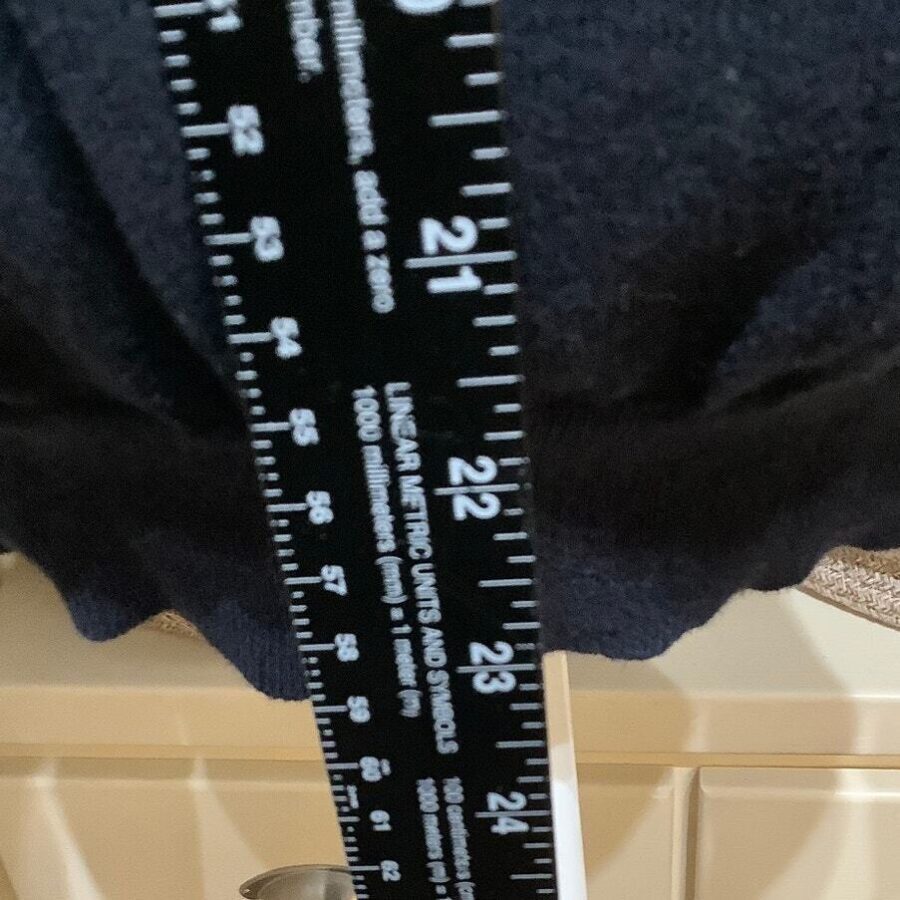 A black measuring tape displaying inch and centimeter markings is held against a dark fabric in what appears to be an indoor setting.