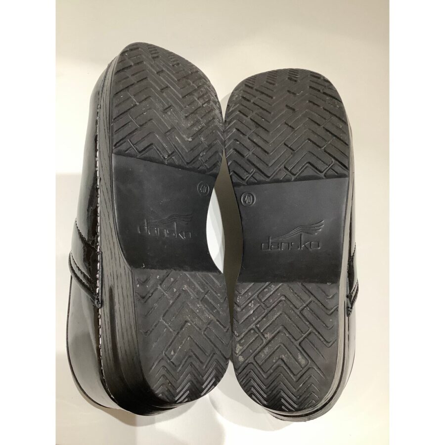 A close-up of the soles of a pair of black Dansko shoes, displaying tread patterns and logo against a white background.