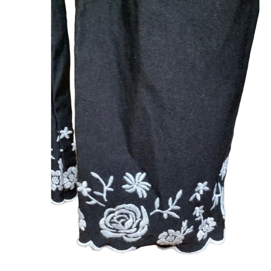 A black fabric with intricate white floral embroidery along the hemline, featuring various flowers and scalloped edges, displayed against a plain background.