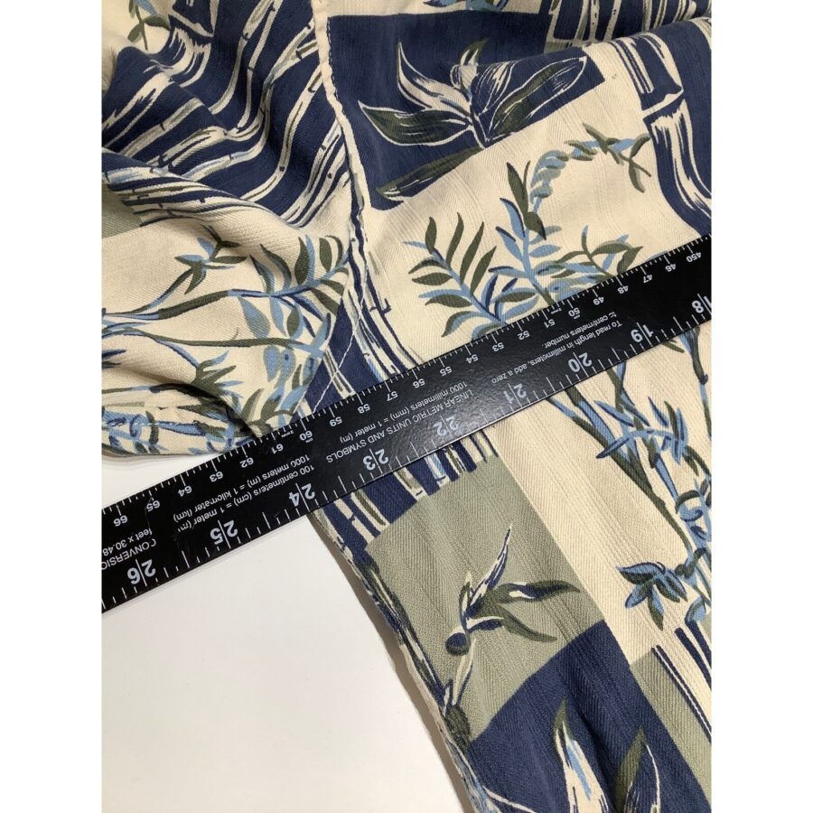 A patterned fabric with green leaves and blue accents is draped, with a black ruler placed diagonally on top, measuring in both inches and centimeters.