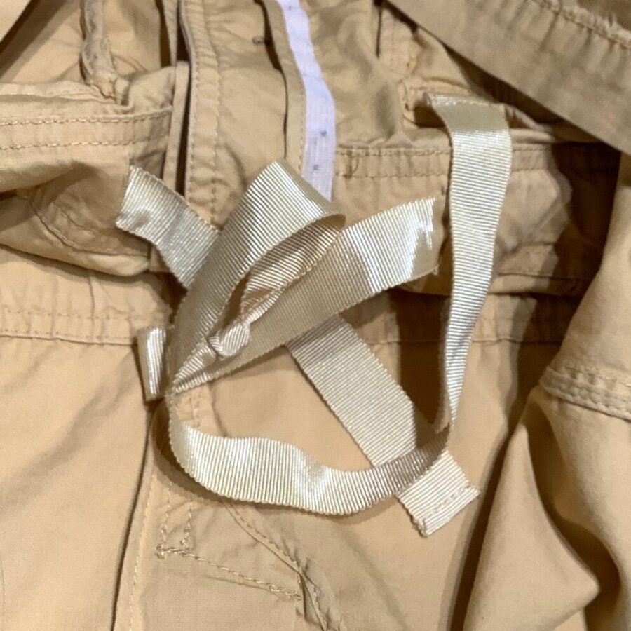 The image shows a close-up of beige clothing with cream-colored ribbons tied in a knot. The fabric appears to be part of a jacket.