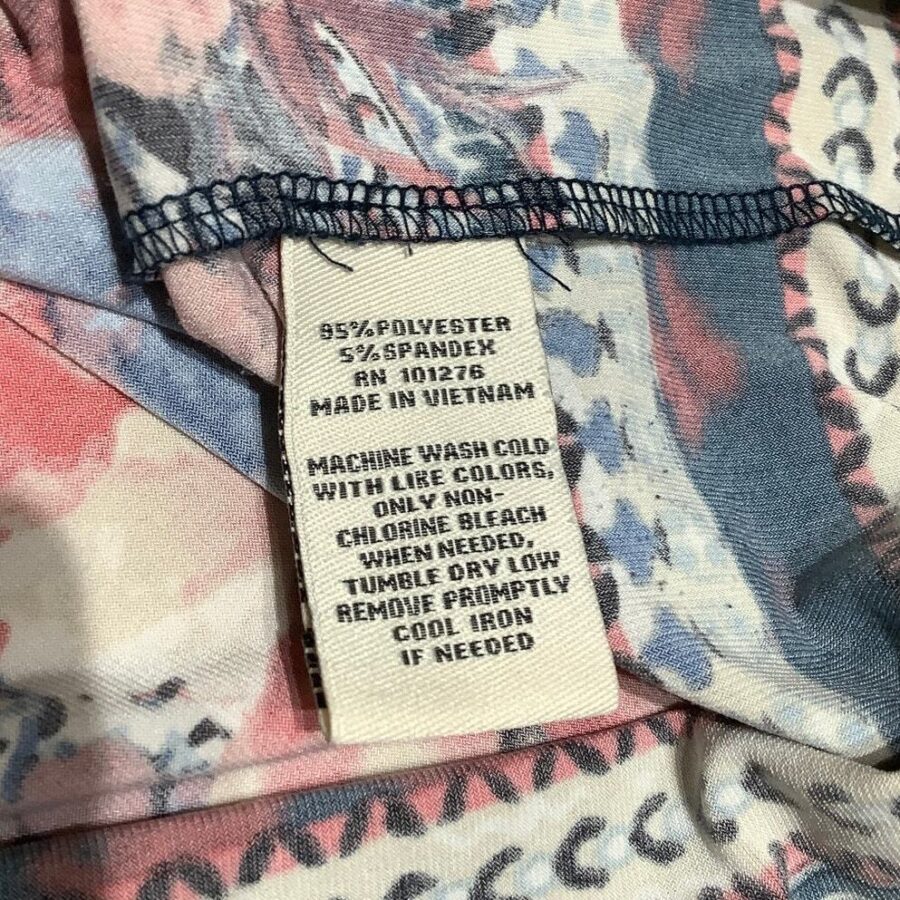 Close-up of a garment's care label indicates material composition, country of origin, and washing instructions. Background shows a multicolor fabric pattern.