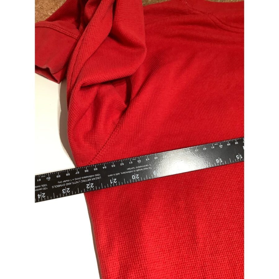 A red shirt is shown laid flat with a black ruler measuring its width from armpit to armpit. No landmarks or historical buildings visible.