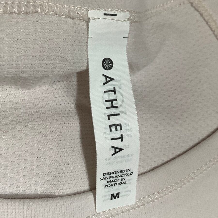 Close-up of a clothing tag on a beige garment. Tag reads "ATHLETA," "Designed in San Francisco," and "Made in Portugal," with a size "M" label.