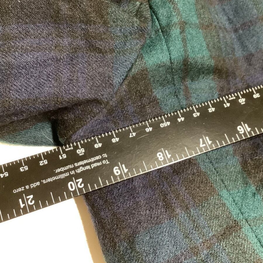 A close-up of a measuring tape placed on a piece of dark plaid fabric, showing centimeters and inches for measurement.