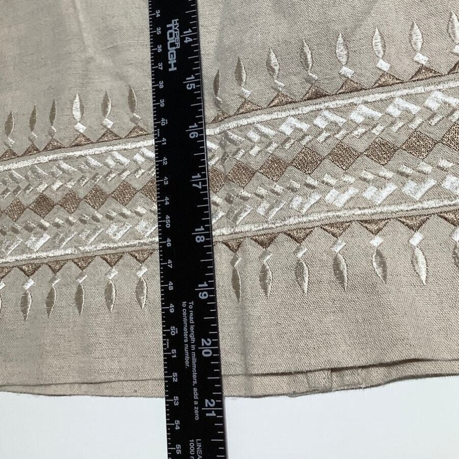 A close-up of a fabric with intricate white and beige embroidery, alongside a black ruler for scale, displaying measurements in inches and centimeters.