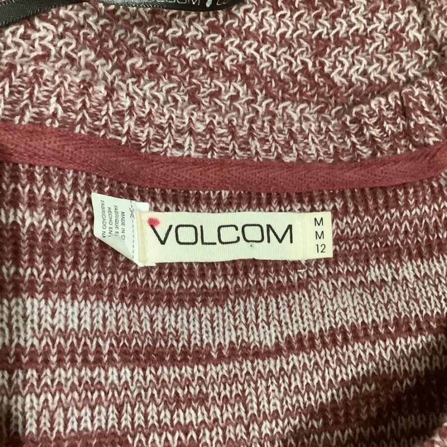 The image shows a close-up of a red and white knitted Volcom sweater, displaying a clothing tag with sizing information. No landmarks or historical buildings are visible.