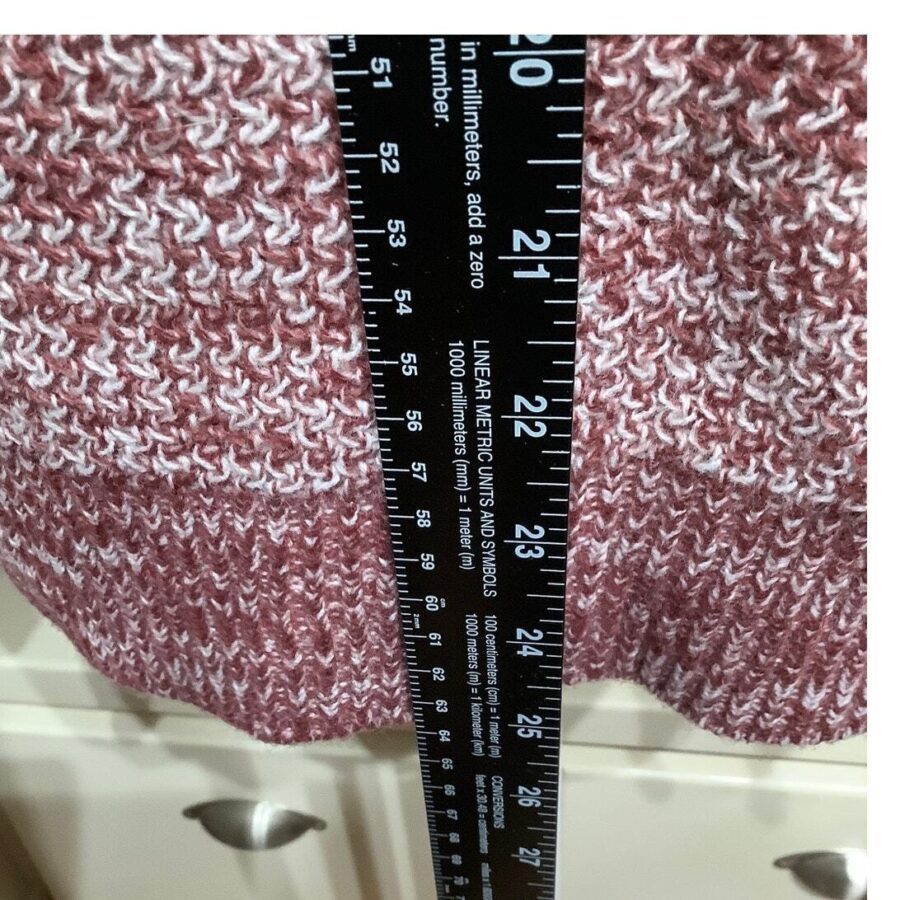 A black ruler measures a pink and white knitted fabric. The fabric is held over a white dresser drawer with round knobs.