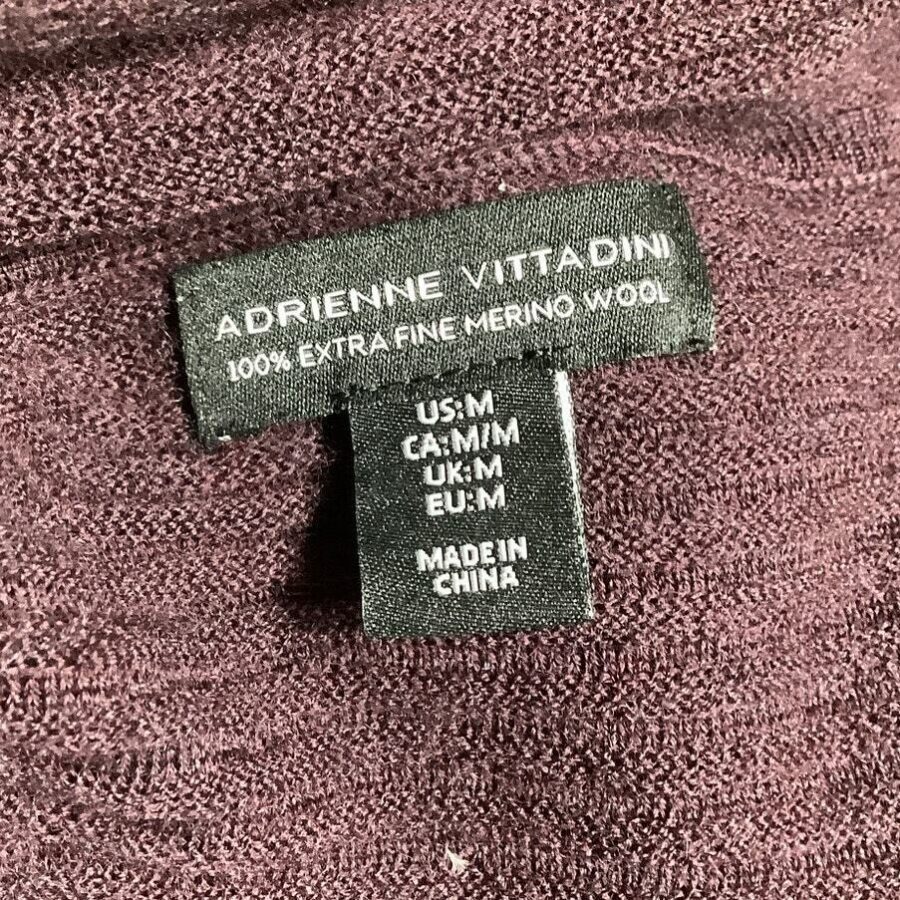 A clothing label with "Adrienne Vittadini," indicating a medium size (M) in several regions and made from extra fine Merino wool, made in China.