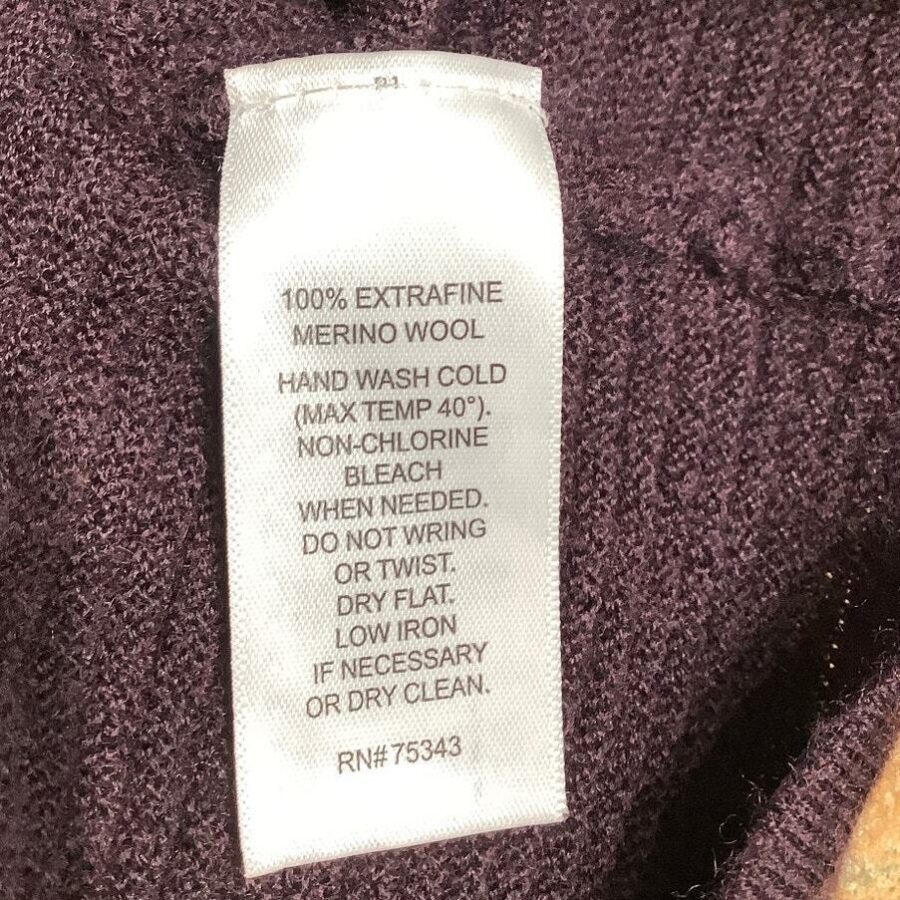 Clothing care label on purple fabric. Instructions for washing and handling 100% extrafine merino wool, including hand wash, non-chlorine bleach, low iron, or dry clean.