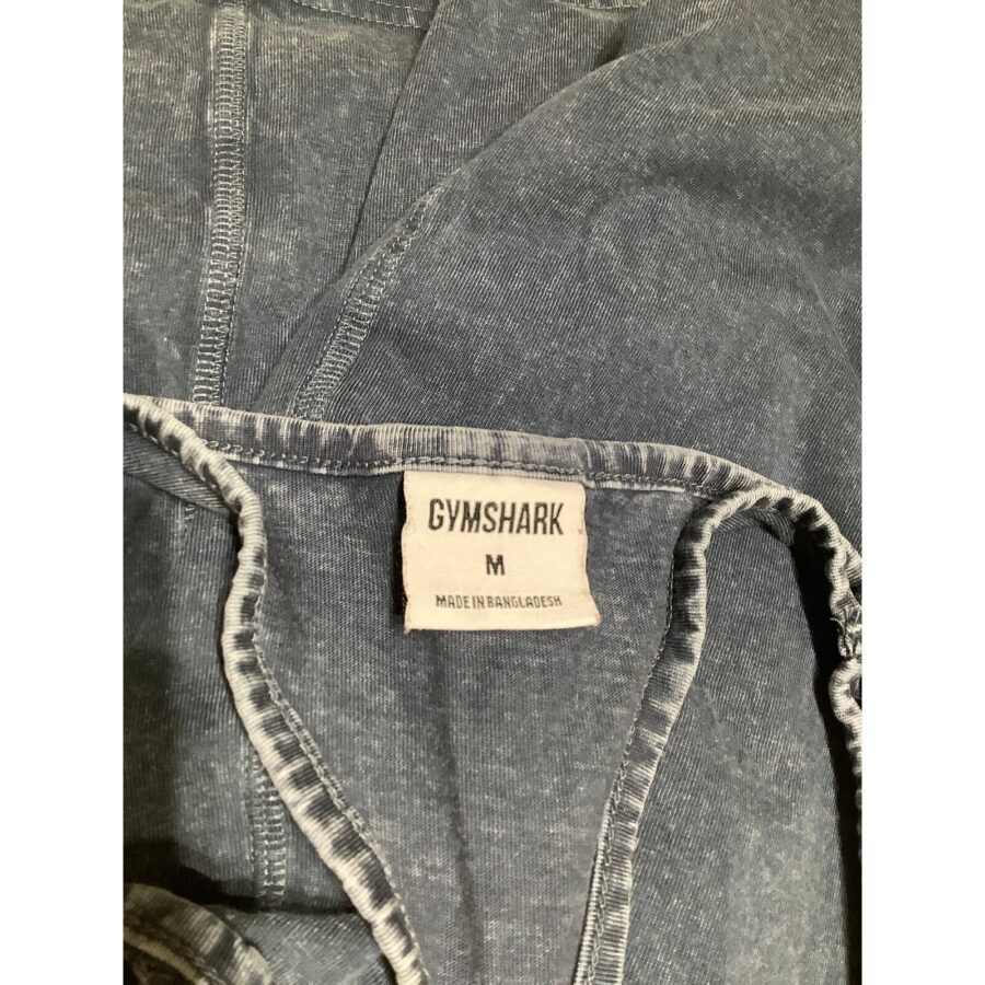 A Gymshark clothing label is visible on a dark fabric with stitching details. The label indicates the size M and origin, Bangladesh.